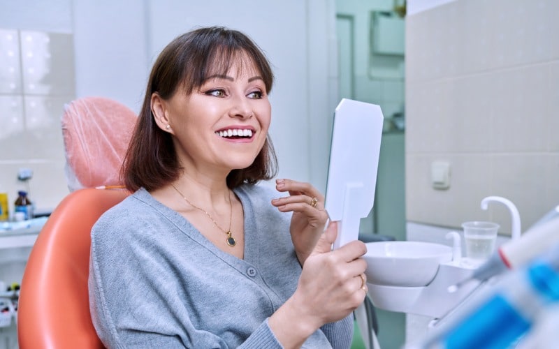 smiling middle aged woman in dental chair with mirror looking at her teeth
